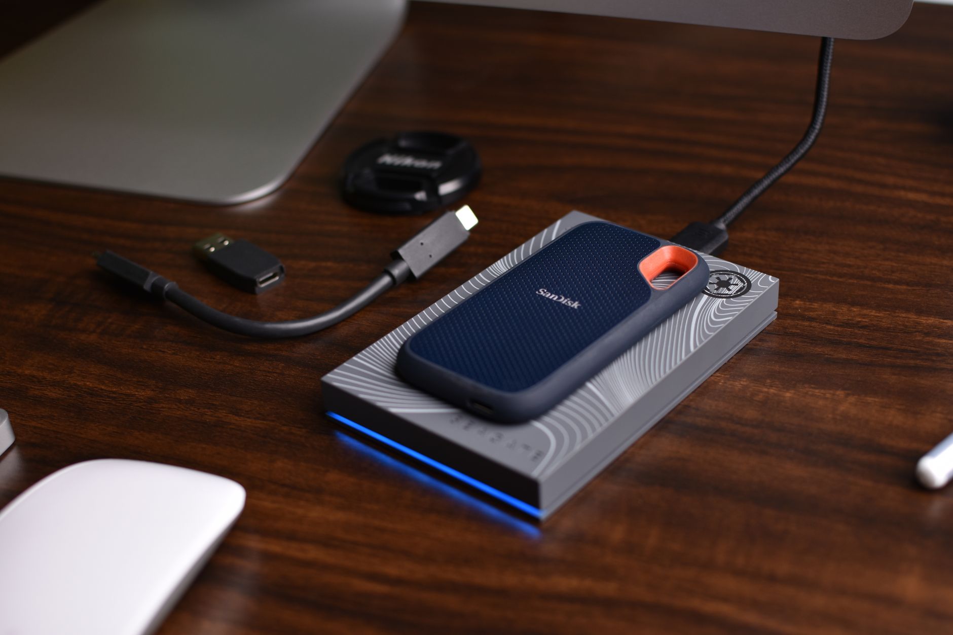 SSD portable SanDisk posé sur un disque dur externe gris, avec câble USB-C et adaptateur, sur un bureau en bois.